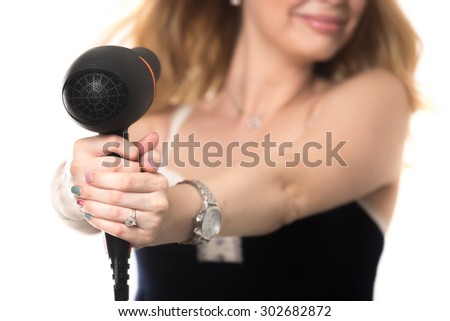Young attractive blond woman drying her hair, holding black hairdryer on stretched arms pointing at her face, fooling around, close up, focus on dryer, studio shot, isolated on white background