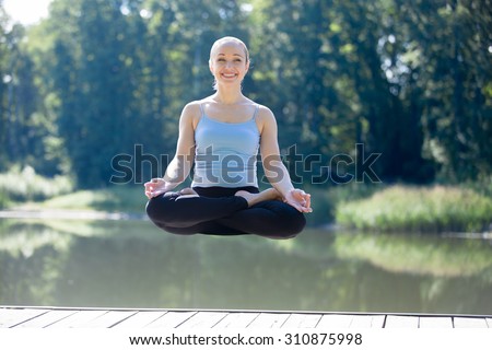 Portrait of happy smiling young beautiful woman meditating in midair, floating outdoors above ground, working out in park on summer day, sitting with crossed legs in Lotus Posture, Padmasana