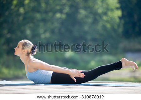 Beautiful sporty fit young woman in sportswear working out outdoors in park on summer day, doing Salabhasana, Locust Posture, Double Leg Kicks exercise, backward extension of the spine, full length