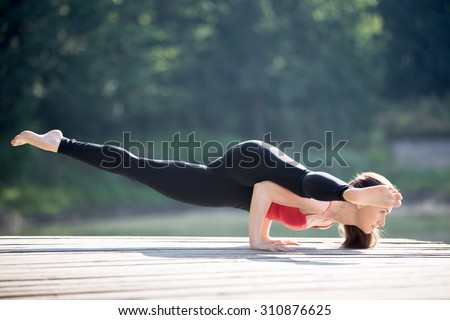 Fit young beautiful woman wearing red tank top and black sporty leggings working out outdoors in park on summer day, doing Eka Pada Koundinyasana 2, twisted one legged arm balance
