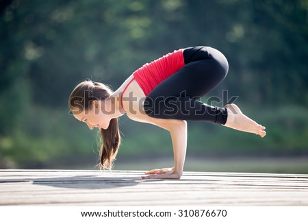 Fit young beautiful smiling woman wearing red tank top and black sporty leggings working out outdoors in park on summer day, doing Crane (Crow) Pose, Bakasana (Kakasana), full length