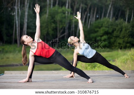 Two fit young beautiful women doing utthita parshvakonasana (Extended Side Angle posture), working out outdoors in park on summer day, wearing sportswear red and blue tank tops, full length
