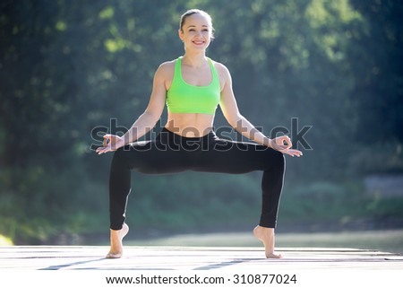 Beautiful sporty fit smiling blond young woman working out outdoors on wooden deck, standing in Goddess (Fierce Angle Pose) or sumo squat posture, full length
