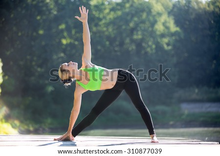 Beautiful sporty fit blond young woman in green sportswear working out outdoors on wooden deck, standing in Utthita Trikonasana, Extended Triangle posture, full length