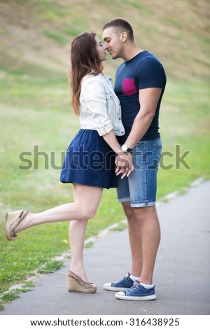 Happy beautiful young couple on a date standing close to each other in park, about to kiss, holding hands, flirting, girl playfully lifted her leg trying to reach his lips