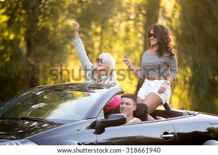 Group of three carefree smiling friends going on trip, traveling together in car on sunny summer day, girls having fun, rising hands, laughing