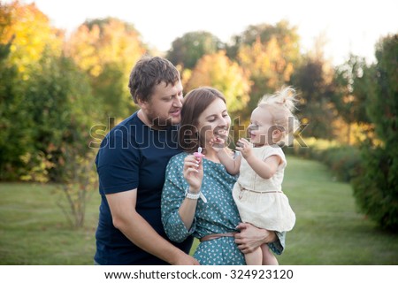 Portrait of happy beautiful family of three people walking in park in autumn. Mom and dad carrying baby daughter in white dress, playing, trying to take away pacifier and laughing