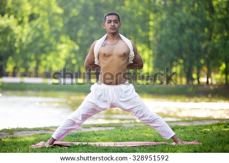 Attractive Indian young man in white linen clothes working out on riverbank in park, doing preparation for Pyramid Pose, Intense Side Stretch Posture, Parsvottanasana, hands behind back, full length
