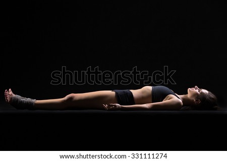 Beautiful cool young fit woman in sportswear lying in Shavasana (Savasana, Corpse or Dead Body Posture), resting after practice, meditating, breathing, side view, studio, black background