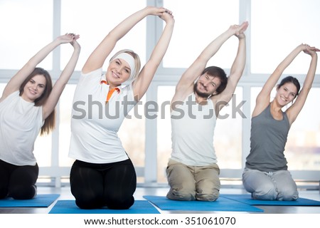Fitness, stretching practice, group of four beautiful happy fit young people working out in sports club, doing side bend exercises with raised arms on blue mats in class