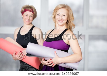 Indoor portrait of group of two cheerful attractive fit senior women posing holding fitness mats, working out in sports club class, happy smiling, looking at camera with friendly expression