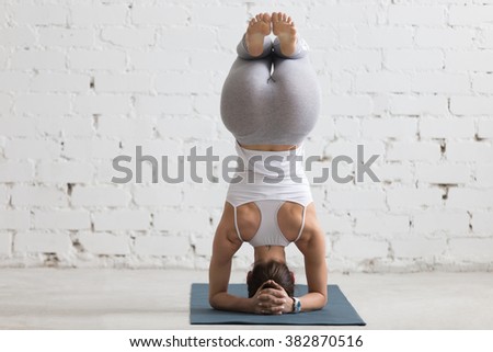 Beautiful young woman with sports smart watch working out indoors, doing yoga exercise on blue mat, variation of supported headstand, salamba sirsasana with bent legs, back view, full length