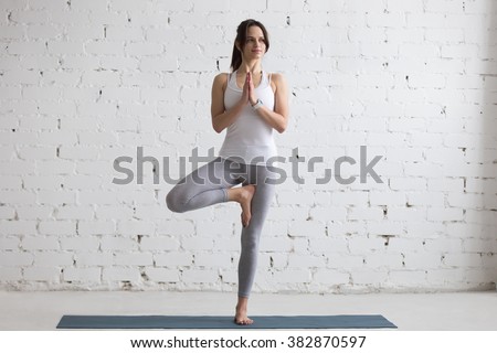 Beautiful smiling happy young woman working out indoors, doing yoga exercise on blue mat, standing in Vrksasana Posture, Tree Pose, full length