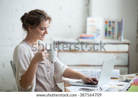 Portrait of beautiful happy smiling young designer woman sitting at home office desk with cup of coffee, working on laptop in loft interior. Attractive cheerful model using computer, typing. Indoors