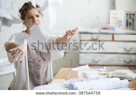 Young stressed woman sitting at desk in a little office or home mad at work, ripping documents with frustrated facial expression. Throwing around scraps of paper. Negative human emotions