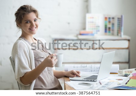 Portrait of beautiful happy smiling young designer woman sitting at home office desk with cup of coffee, posing, looking at camera. Attractive cheerful model using computer. Indoors