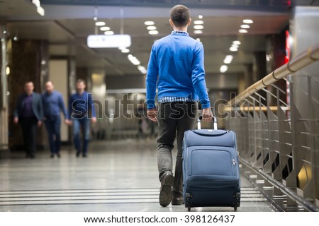 Young man pulling suitcase in modern airport terminal. Travelling guy wearing smart casual style clothes walking away with his luggage while waiting for transport. Rear view. Copy space