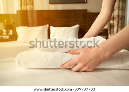 Close-up of hands putting stack of fresh white bath towels on the bed sheet. Room service maid cleaning hotel room. Lens flair in sunlight