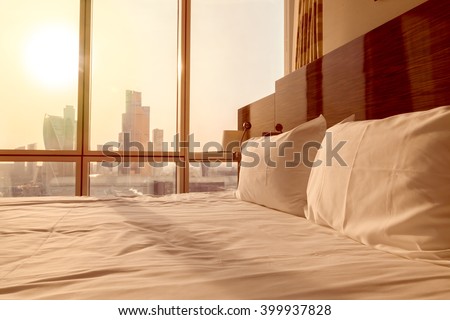 Bed maid-up with clean white pillows and bed sheets in empty room. Close-up. Lens flair in sunlight. Sunrise city view on the background