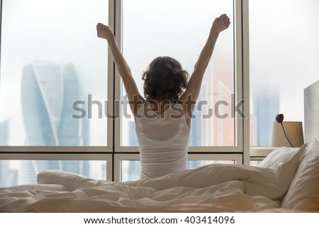 Back view of young woman stretching on unmade bed after waking up and looking at city view in the window. Female model relaxing at home in the morning. Motivation concept