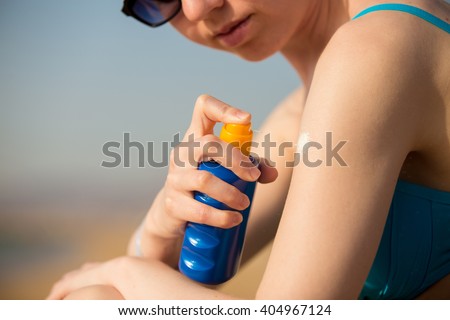 Young woman in sunglasses holding bottle of sunscreen lotion, spraying sunblock cream on shoulder before tanning, close up