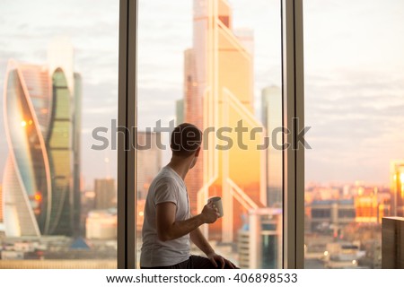 Back view of young man sitting on window with coffee cup, looking at dawn city scenery after early wakeup. Handsome casual guy relaxing in penthouse after good night sleep and watching sunrise