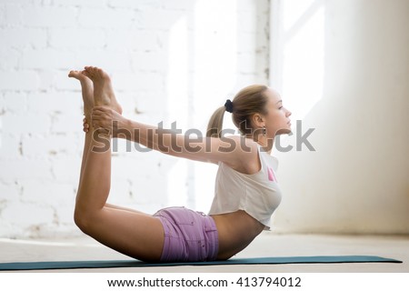 Beautiful young woman wearing casual clothing enjoying yoga indoors. Yogi girl working out in sunny loft interior. Doing Dhanurasana, Bow Pose. Full length