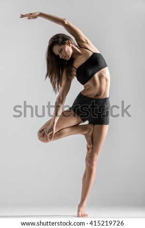 Portrait of beautiful young woman wearing black sportswear working out in studio. Fit sporty girl doing side bend exercise. Variation of Vrksasana Posture, Tree Pose. Full length. Vertical image