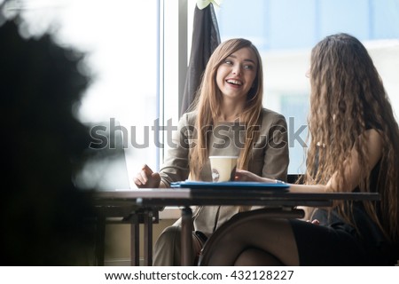 Portrait of two beautiful young girlfriends sitting in modern coffee shop interior and talking with happy smiles. Successful attractive women friends chatting in cafe during coffee break