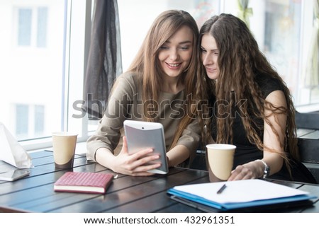 Portrait of two beautiful young girlfriends using app on tablet computer in modern coffee shop interior. Successful attractive women friends looking at tablet screen with happy expressions indoors