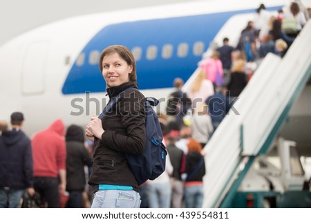 Young Caucasian cheerful smiling woman passenger in 20s travelling with backpack, boarding airplane, people climbing ramp on background