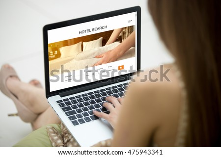 Casual young woman sitting on sofa with notebook, working on laptop computer, searching for lodging using on-line web service, booking a hotel on website. Closeup view over the shoulder