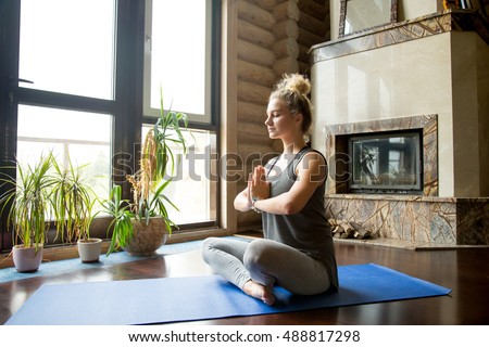 Full length portrait of attractive young woman working out at home, doing yoga exercise on blue mat, sitting in Easy (Decent, Pleasant Posture) with palms in Namaste, meditating, breathing, relaxing
