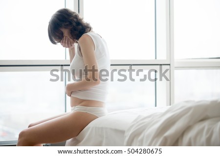 Profile view of young pregnant woman sitting on the bed covering her mouth feeling nauseous in early stages of pregnancy. Morning sickness
