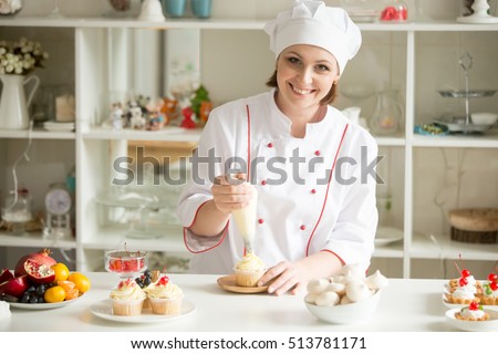 Portrait of friendly smiling female professional confectioner topping a cupcake with cream using a pastry bag. Looking at the camera. Indoors image. Pastry chef woman making creamy cakes