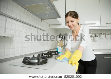 Young smiling woman wearing rubber protective yellow gloves cleaning the stove with a rag and spray bottle detergent. Home, housekeeping concept. Looking at the camera