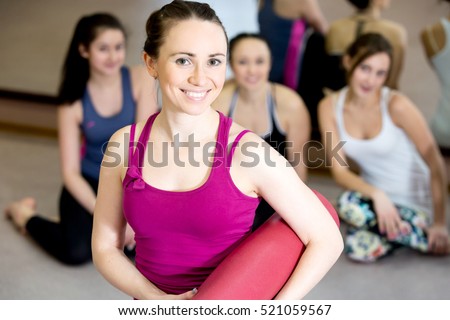 Portrait of beautiful happy yogi girl wearing bright tank top is holding folded pink yoga mat after training lesson in fitness center or gym, her class partners sitting on the background