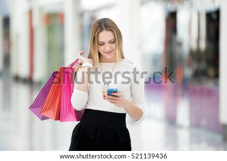Good news and purchases. Happy teenage girl holding bags with purchases, smiling while looking at phone in shopping center. Reading message, texting, dialing number, using app on smartphone