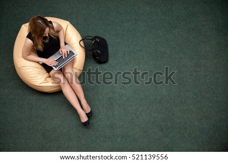 Young beautiful woman sitting on bean bag working on laptop in public wifi area, typing, top view, copy space. Green carpet on the background floor. Full length