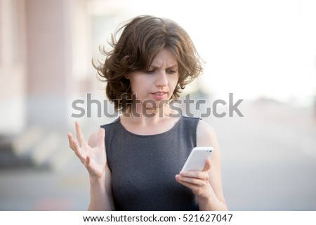 Portrait of young angry woman holding cellphone in hands on the street in summer, looking at screen with irritated expression, made a mistake or annoyed by texts and calls