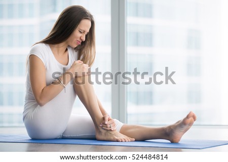 Unhappy young woman sitting on the mat, grabbing an ankle, unable to start yoga work out because of sport injury, feeling pain. Beginner doing wrong exercise without coacher