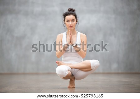 Young calm pretty woman wearing white sportswear working out against grey wall, doing yoga or pilates exercise. Sitting in squat position, Half Lotus Toe Balance, variation of Utkatasana. Full length