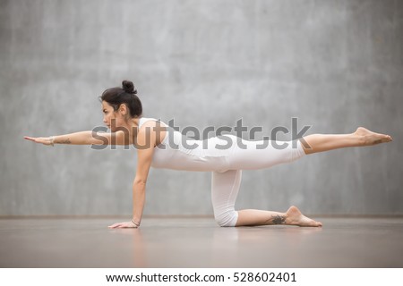 Side view portrait of attractive young woman with beautiful tattoo working out against grey wall, doing yoga, pilates balancing exercise. Bird dog, chakravakasana pose. Full length