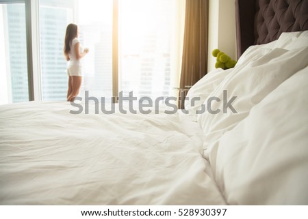 Close up of a made bed with white quality bed linen, bedding, staples quilts pillows, woman standing near the window, welcoming a new day, having cup of coffee, seeing the husband off