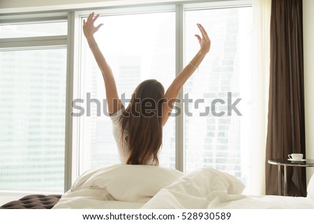 Young woman outstretching her arms sitting on the bed after good night sleep, unwilling to get up and leave her comfortable nest, entering a day happy and relaxed, ready for productive work. Rear view