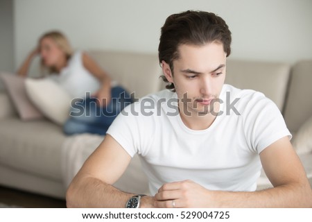 Troubled young handsome man, woman at the background on the sofa, sex life is nonexistent, no displays of physical affection between both, becoming strangers. Family relationship problems concept