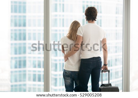 Rear view of attractive young couple in modern apartment looking in full length window at big city scenery, holding suitcase, leaving hometown, or arriving to new place, travelling together. Indoors