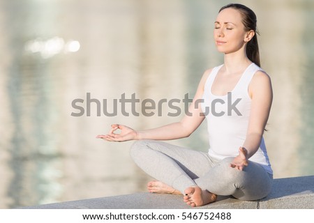 Sporty beautiful young woman practicing yoga, sitting in Easy Seat exercise, Sukhasana pose, working out, enjoying meditation session. Outdoor full length, water surface background. Copy space