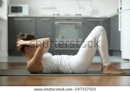 Sporty young attractive woman practicing abdominal, crunches, sit ups, rock press exercise, working out, wearing white sportswear, indoor full length, home interior background