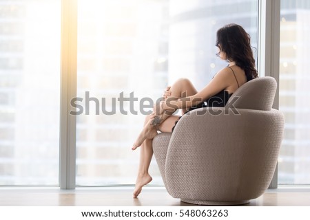Back view of young barefoot model woman sitting in modern armchair, looking at city after waking up at morning, spend time relaxing, relaxation before bedtime, after long day. Skincare, beauty concept